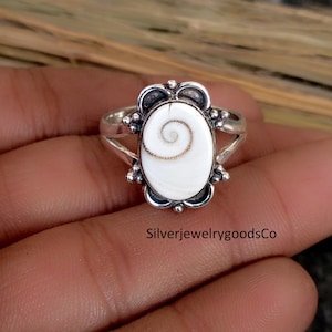 Puede incluir: Anillo de plata con una piedra ovalada blanca con un diseño en espiral. El anillo tiene un engaste ornamentado con pequeños detalles redondos. El texto "SilverjewelrygoodsCo" es visible en la parte inferior.