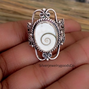 Puede incluir: Anillo de plata con una piedra ovalada blanca con un diseño en espiral. El anillo tiene un diseño de filigrana ornamentado alrededor de la piedra y en la banda. El texto "SilverjewelrygoodsCo" es visible.