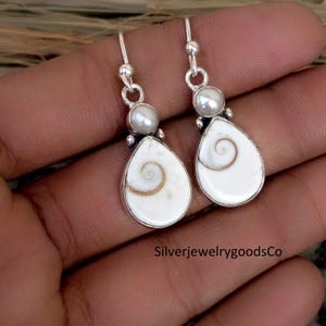 May include: A pair of silver earrings with a pearlescent white teardrop-shaped pendant. Each earring features a small, round pearl at the top and a spiral design within the pendant. The text "SilverjewelrygoodsCo" is visible.