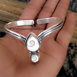 Puede incluir: Una pulsera plateada con un colgante blanco en forma de espiral. La pulsera tiene un diseño texturizado a lo largo de la banda y una pieza central en forma de lágrima con un colgante más pequeño y a juego debajo. El texto "SilverjewelrygoodsCo" es visible.