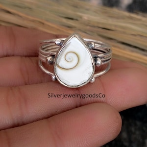 Pode incluir: Um anel de prata com uma pedra branca em forma de gota com um desenho em espiral. O anel tem várias faixas e pequenas contas de prata. O texto "SilverjewelrygoodsCo" é visível na parte inferior da imagem.