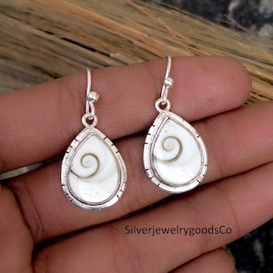 Puede incluir: Un par de pendientes de plata con un diseño en forma de lágrima. Cada pendiente presenta una incrustación de concha blanca con remolinos dentro de un marco plateado. Los pendientes tienen un cierre de gancho simple. El texto "SilverjewelrygoodsCo" es visible en la parte inferior.