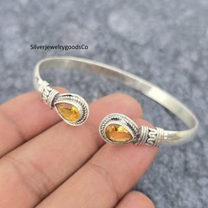 Può includere: Bracciale a polsino in argento con due gemme gialle a forma di goccia. Il bracciale aperto presenta dettagli decorativi attorno alle gemme. Il testo "SilverjewelrygoodsCo" è visibile.