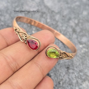 May include: Open cuff bracelet with a copper finish, featuring a red teardrop-shaped gemstone on one end and a green gemstone on the other. The bracelet has decorative filigree details near the gemstones.