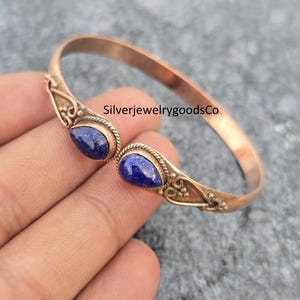 Puede incluir: Una pulsera brazalete de color cobre con dos piedras preciosas azul oscuro en forma de lágrima. La pulsera tiene detalles ornamentados alrededor de las piedras preciosas y en los lados. El texto "SilverjewelrygoodsCo" es visible.