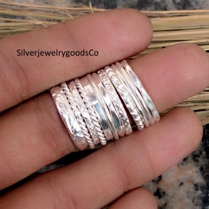 Può includere: Una collezione di anelli d'argento in vari design, tra cui fasce intrecciate, floreali e semplici. Gli anelli sono impilati su un dito, mostrando le loro diverse trame e stili. Gli anelli sono in argento lucido. Il testo "SilverjewelrygoodsCo" è visibile.