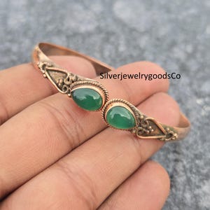 Puede incluir: Una pulsera de color cobre con dos piedras preciosas ovaladas de color verde esmeralda. La pulsera tiene un diseño ornamentado con el texto "SilverjewelrygoodsCo". La pulsera se sostiene en una mano.