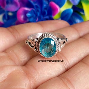 May include: A silver ring featuring a central oval-cut blue gemstone surrounded by a twisted rope-style metal border. The band displays decorative scrollwork details on the sides. The text SilverjewelrygoodsCo is visible on the ring surface.