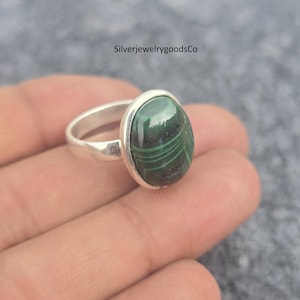 May include: A silver ring with a large, oval malachite gemstone. The malachite is a rich green with darker green patterns. The ring's band is plain, polished silver. The ring is shown against a neutral backdrop.