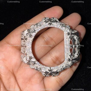 Può includere: Una cassa per orologio argentata con un design a croce e gemme. La cassa ha un'apertura circolare ed è incastonata con piccole pietre scintillanti. Il testo "Customebliing" è visibile.