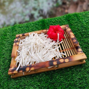 May include: A bamboo tray with white, matchstick-cut vegetables and a bright red hibiscus flower. The tray has a natural wood finish with dark brown accents. The tray is set on green artificial grass.