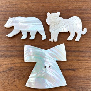 May include: Three decorative items made of iridescent mother-of-pearl. Two pieces are carved into the shapes of a bear and a tiger, while the third piece is a flat, flared garment-shaped object featuring two small holes in the centre for attachment.