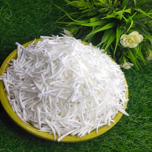 May include: A yellow bowl filled with shredded white vegetables, possibly daikon radish or a similar root vegetable. The bowl sits on a bed of green grass, with green foliage and a white flower in the background.