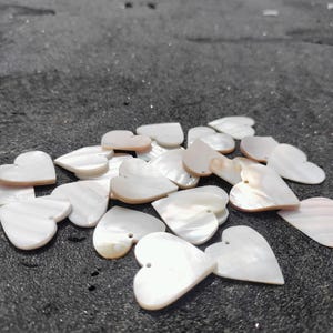 May include: A scattering of white, heart-shaped pendants with a pearlescent sheen, lying on a dark, textured surface. Each pendant has a small hole for stringing. The pendants are likely made of mother-of-pearl.