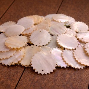 May include: A collection of circular mother of pearl shell components, each featuring a serrated, gear-like edge and two small holes positioned in the centre for attachment. The pieces exhibit a natural, iridescent off-white colour palette.