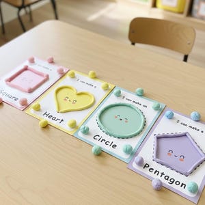 May include: Four educational shape cards labelled "Square", "Heart", "Circle", and "Pentagon". Each card features a 3D shape and small pom-poms. The cards are displayed on a light wood table.