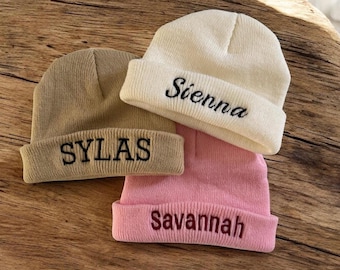 Può includere: Tre berretti in maglia personalizzati in beige, crema e rosa. Ogni berretto ha un nome ricamato in un colore a contrasto: "SYLAS" in nero, "Sienna" in nero e "Savannah" in bordeaux. I berretti sono ripiegati sul bordo.