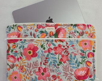 Op de afbeelding: Een gewatteerde laptophoes met een lichtgrijze achtergrond en een levendig bloemenpatroon in roze, rode, oranje en blauwe tinten. De hoes heeft een witte ritssluiting en een klein rechthoekig label met de tekst Sample Photo.