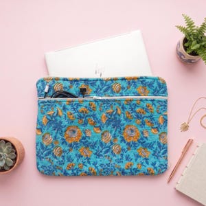 May include: A quilted laptop sleeve featuring a vibrant turquoise background with a repeating pattern of orange and blue floral motifs. The sleeve includes a horizontal zippered pocket across the top, containing black charging cables and a white laptop.