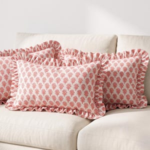 May include: A rectangular throw cushion featuring a repeating coral-red floral block print pattern on a cream background. The cushion is finished with a gathered ruffle trim along all four edges, creating a soft, textured border around the design.
