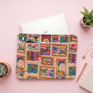 May include: A quilted laptop sleeve featuring a beige background with a repeating pattern of colorful postage stamp-style illustrations, including sailboats, fish, and abstract shapes, with a zippered pocket containing black charging cables.
