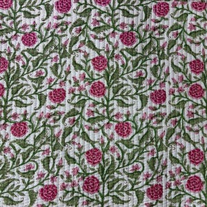 May include: A white quilted fabric featuring a repeating floral pattern with vibrant pink roses and intricate green leafy vines. The textile displays vertical stitching lines throughout, creating a textured surface with a consistent botanical design.