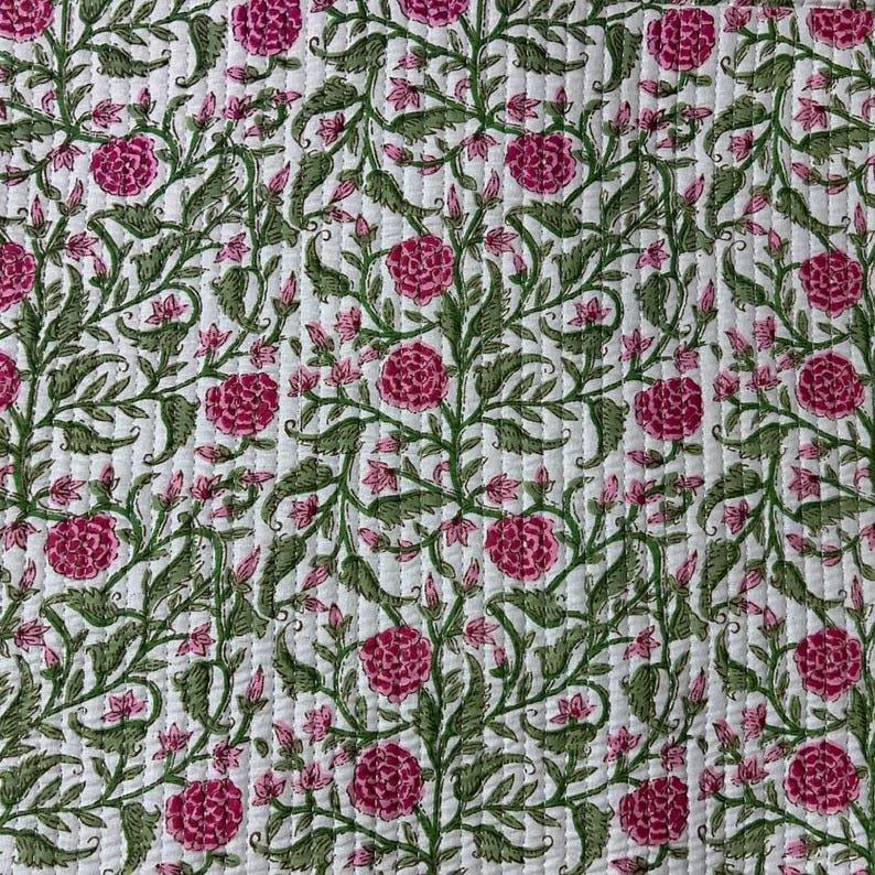 May include: A white quilted fabric featuring a repeating floral pattern with vibrant pink roses and intricate green leafy vines. The textile displays vertical stitching lines throughout, creating a textured surface with a consistent botanical design.