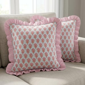 Könnte beinhalten: Zwei dekorative Zierkissen mit einem Blumenmuster in Rosa- und Orangetönen auf hellblauem Hintergrund. Die Kissen haben einen gerüschten Rand und einen rosa-weiß gestreiften Rand. Sie liegen auf einem neutralfarbenen Sofa.