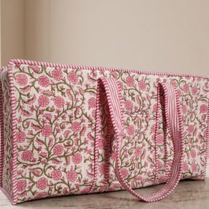 May include: A rectangular quilted tote bag featuring a pink floral pattern on an off-white background. The bag has two matching fabric handles and pink and white striped piping along the edges and straps, providing a structured appearance.