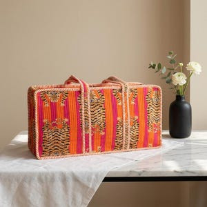 May include: A rectangular quilted tote bag featuring a vibrant pattern of orange and pink vertical stripes with repeating tiger illustrations. The bag has two fabric handles and is finished with a light-colored piped trim along the edges.