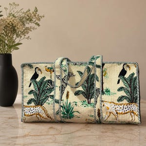 May include: A rectangular quilted tote bag featuring a cream-colored fabric with a repeating jungle print. The pattern includes green palm trees, black and white zebras, spotted cheetahs, and toucans. The bag has two handles and black and white trim.