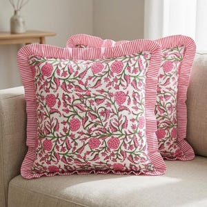 May include: Two square cushions with a floral design. The cushions have a white base with pink flowers and green foliage. The edges are trimmed with a pink and white striped frill.