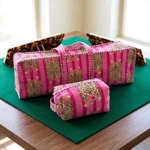 May include: A rectangular quilted bag and a smaller matching zippered pouch, both featuring a vibrant pink and white vertical striped pattern with repeating tiger illustrations. The bags have fabric handles and a quilted texture throughout.