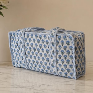 May include: A rectangular quilted fabric storage case featuring a repeating blue floral block print on a white background. The bag has a structured shape with light blue and white striped fabric trim along the edges and two matching fabric handles.