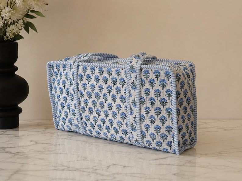 May include: A rectangular quilted fabric storage case featuring a repeating blue floral block print on a white background. The bag has a structured shape with light blue and white striped fabric trim along the edges and two matching fabric handles.