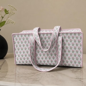 May include: A rectangular quilted fabric storage basket featuring a white background with a repeating pattern of small pink flowers and green leaves, finished with pink and white gingham trim on the edges and two matching fabric handles.