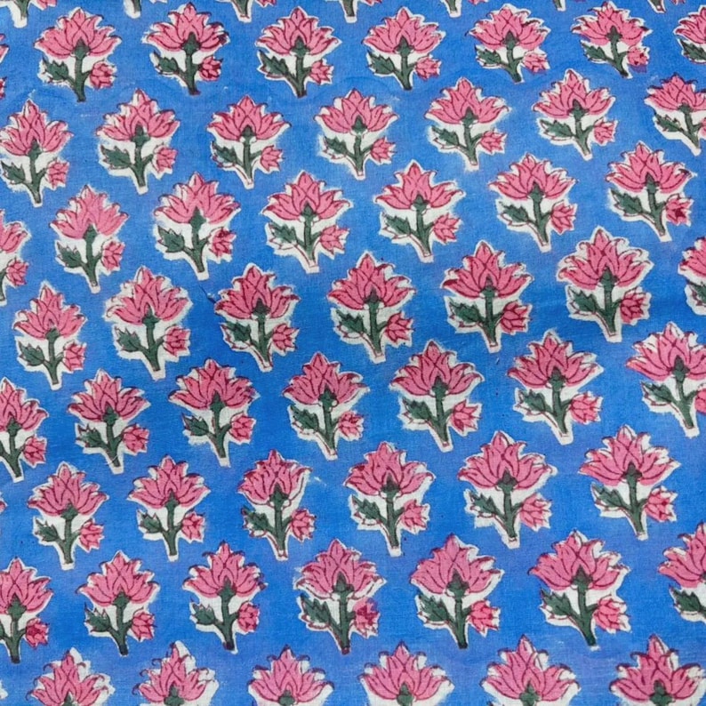 May include: A light blue fabric featuring a repeating pattern of pink lotus flowers with green stems and leaves. The floral design is evenly spaced across the surface, creating a consistent botanical print on the blue textile background.