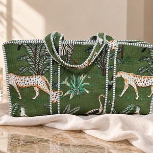 May include: A rectangular quilted tote bag in a deep green fabric featuring a jungle-themed print of cheetahs, zebras, and tropical plants. The bag has two matching fabric handles and is finished with white and dark green striped piping trim.