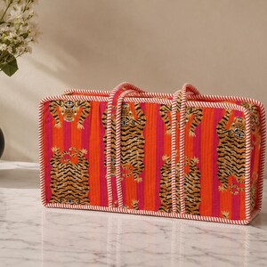 May include: A rectangular quilted tote bag featuring a vibrant pattern of orange and pink vertical stripes with repeating tiger illustrations. The bag is finished with white and red twisted cord piping along the edges and two matching handles.