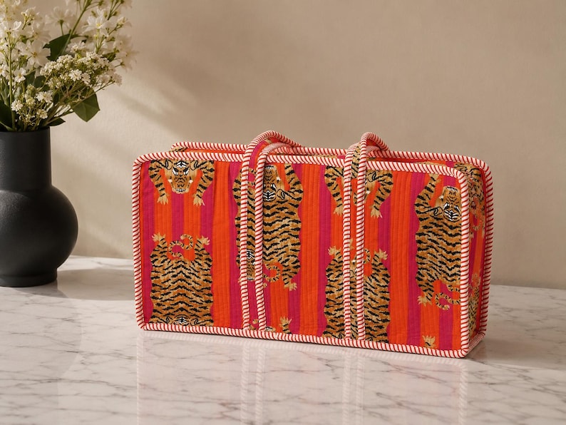 May include: A rectangular quilted tote bag featuring a vibrant pattern of orange and pink vertical stripes with repeating tiger illustrations. The bag is finished with white and red twisted cord piping along the edges and two matching handles.
