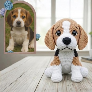 Puede incluir: Un perro beagle de ganchillo hecho a mano con detalles marrones, blancos y negros. El peluche tiene ojos grandes y expresivos y orejas caídas. Una foto enmarcada de un perro beagle real está en el fondo.