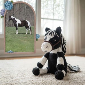 Puede incluir: Un juguete de caballo de ganchillo hecho a mano en blanco y negro, con una brida marrón, está sentado en una alfombra de color crema. El caballo de juguete tiene una cara sonriente y una larga melena y cola blanca y negra. Una foto enmarcada de un caballo real está en el fondo.