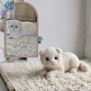 Pode incluir: Um brinquedo de gato de pelúcia creme com olhos azuis e nariz rosa, deitado em um tapete creme. O brinquedo é uma réplica de um gatinho Scottish Fold. Um espelho com reflexo de gato e detalhes florais está ao fundo.