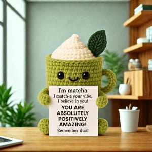 Puede incluir: Una taza de ganchillo con temática de matcha con una cara sonriente, sosteniendo un letrero con afirmaciones positivas. La taza es verde con una cobertura de color crema y una hoja verde. El letrero dice: "I'm matcha, I match-a your vibe, I believe in you! YOU ARE ABSOLUTELY POSITIVELY AMAZING! Remember that!"