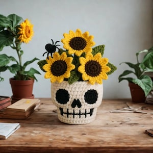 May include: A crocheted skull-shaped planter filled with yellow sunflowers and green leaves. A small black spider sits on top of the flowers. The planter is beige with black eye sockets and a mouth. The sunflowers have brown centers.