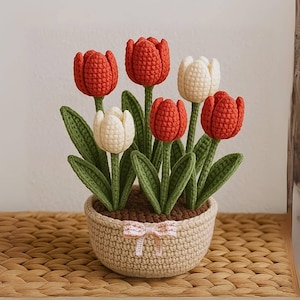 May include: A handcrafted crochet tulip arrangement in a woven basket. The arrangement features red and white tulips with green stems and leaves. The basket is beige with a pink bow. A charming, handmade floral decoration.