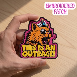 Könnte beinhalten: Gestickter Aufnäher mit einer orangefarbenen Katze, die eine Sonnenbrille und eine Krone trägt, mit dem Text "THIS IS AN OUTRAGE!" in Gelb. Der Aufnäher hat einen rosa Rand und wird von einer Hand gehalten.