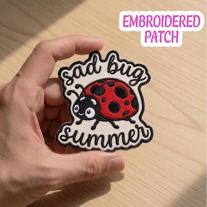 May include: Embroidered patch featuring a cartoon ladybug with a red shell and black spots. The patch has the words "sad bug summer" in a whimsical font. The patch is white with a black border and the text "EMBROIDERED PATCH".