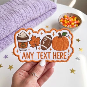 May include: An embroidered patch with a scalloped orange border featuring a coffee cup, a maple leaf, a football, and a pumpkin. The text ANY TEXT HERE is stitched in orange below the icons, accented by small orange stars on a white background.