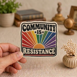 May include: A square embroidered patch with a cream-colored border featuring a rainbow sunburst design. The black text reads COMMUNITY IS RESISTANCE, with the word COMMUNITY arched at the top and RESISTANCE in a straight line at the bottom.